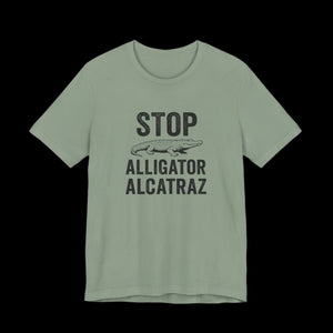 Sage green t-shirt with black text and alligator graphic that says "Stop Alligator Alcatraz" on a black background